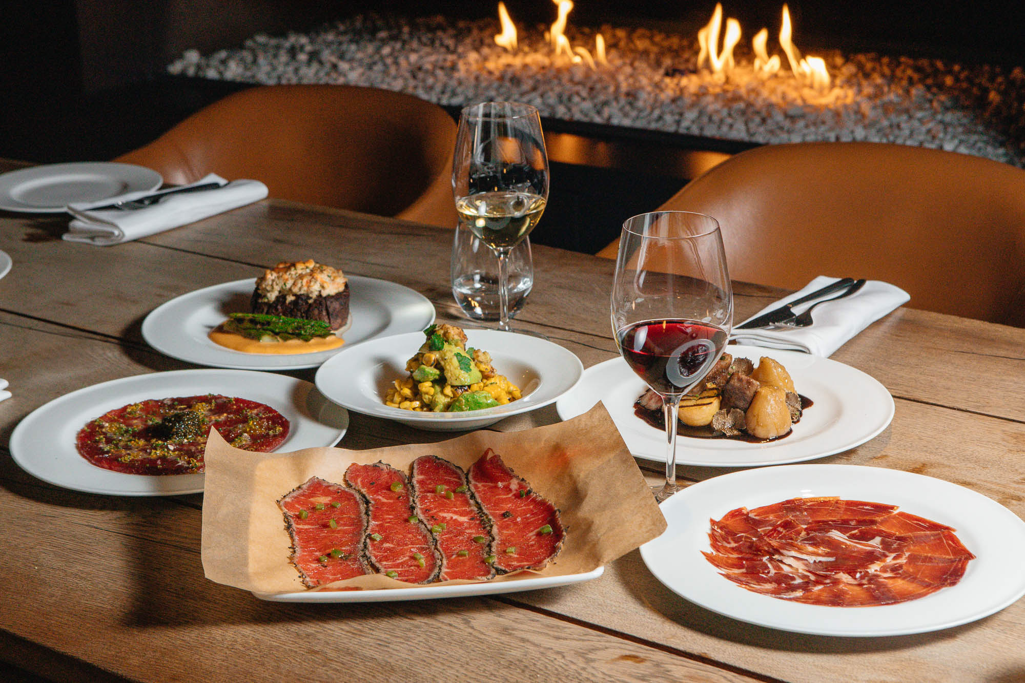 Mesa con platos para compartir y copas de vino frente a una chimenea en MR PORTER.
