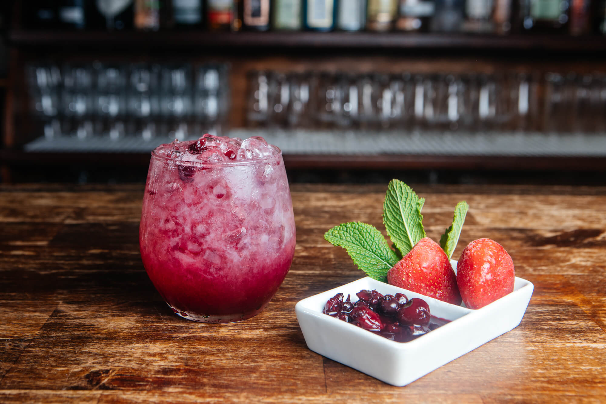 Cóctel rojo con hielo acompañado de fresas, arándanos y hojas de menta.
