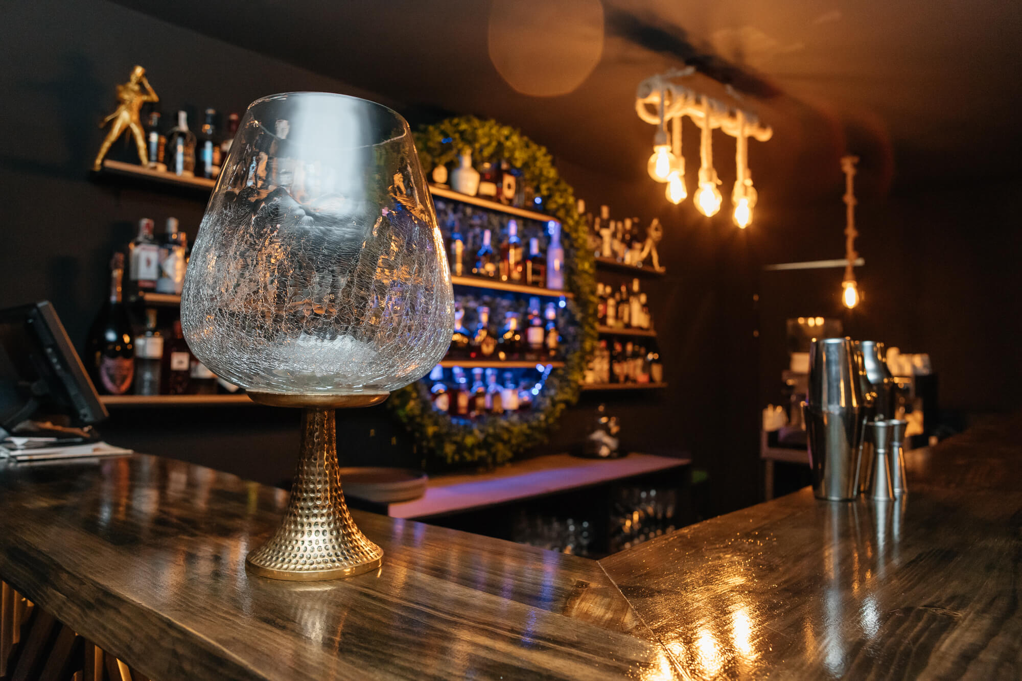 Copa sobre una barra de madera con botellas e iluminación cálida al fondo.