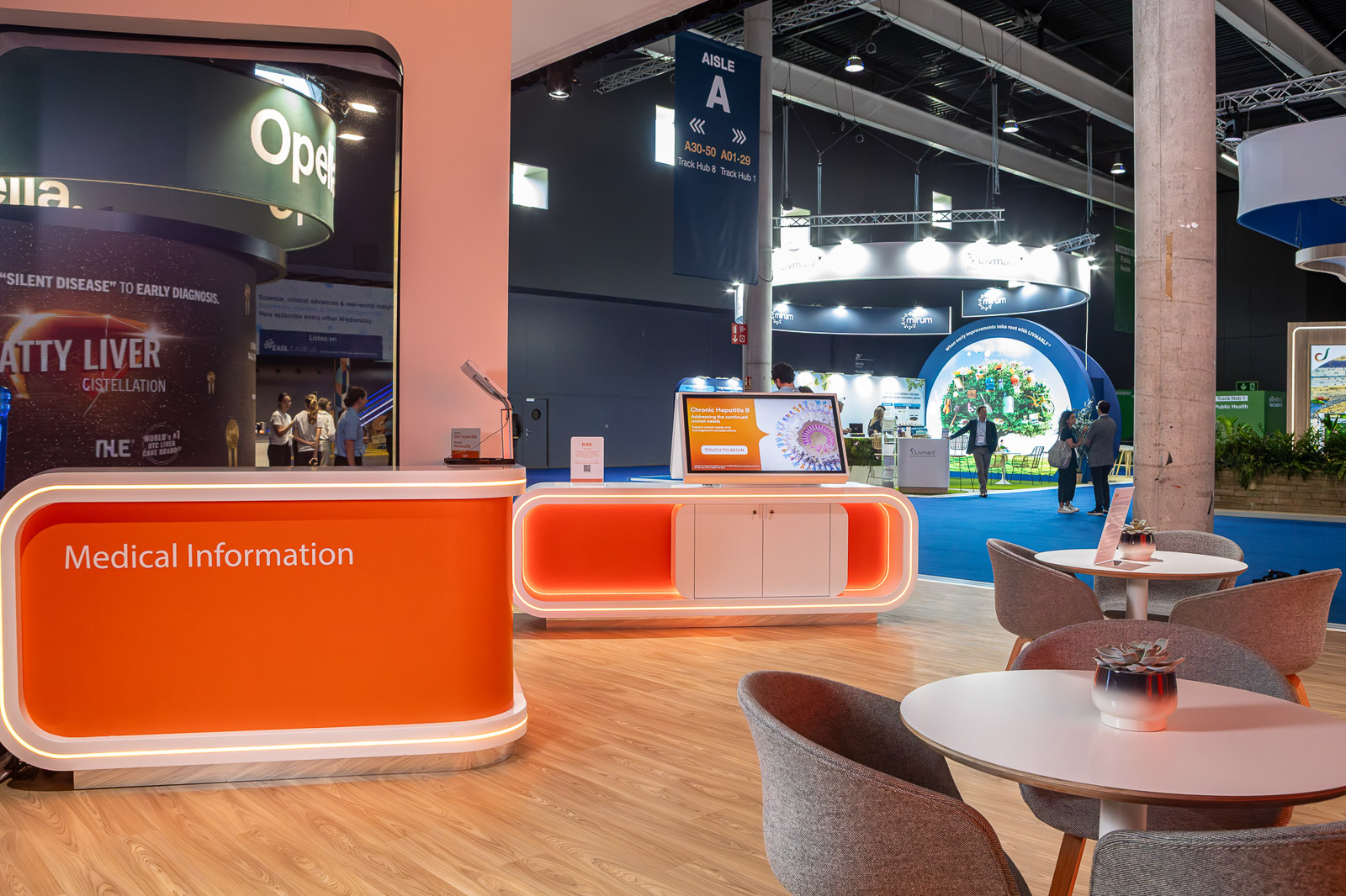 Mostradores de información con iluminación naranja y zona de asientos en el espacio expositivo de GSK.