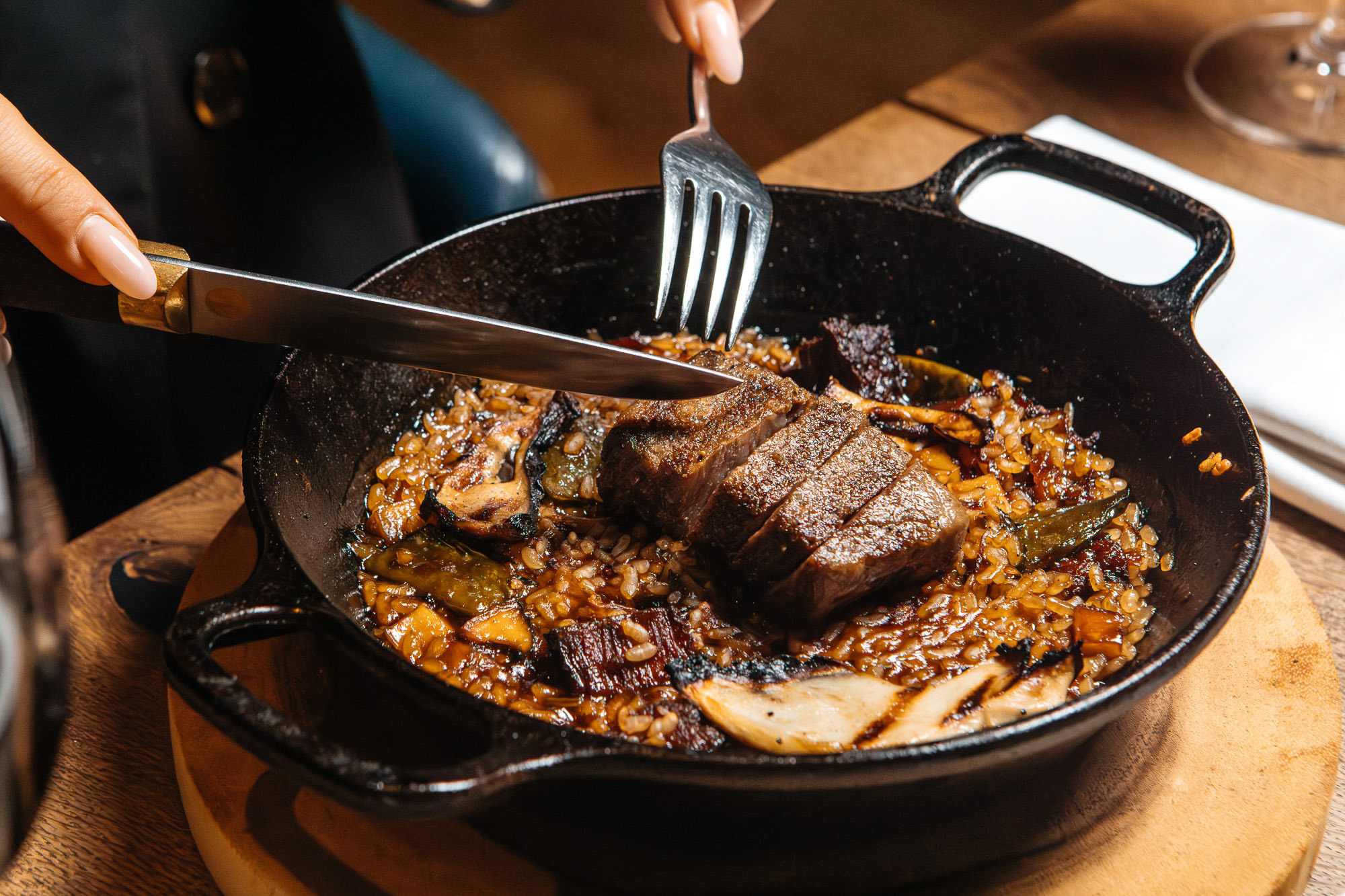Manos cortando carne a la parrilla servida en una sartén con guarnición.